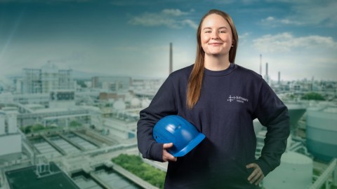 Lächelnde junge Frau mit blauen Helm in der Hand steh vor dem Hintergrund eines Industrieparks.