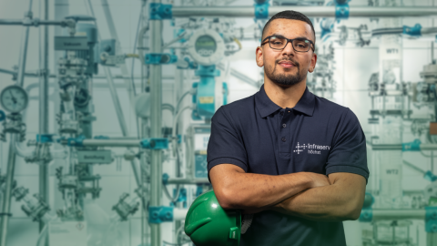 Ein junger Mann mit Brille und blauem Polohemd steht vor einem Hintergrund aus Industrieanlagen. 