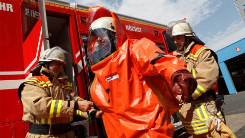 Zwei Feuerwehrleute helfen einem Kollegen in einem orangenen Schutzanzug aus einem Feuerwehrauto auszusteigen.