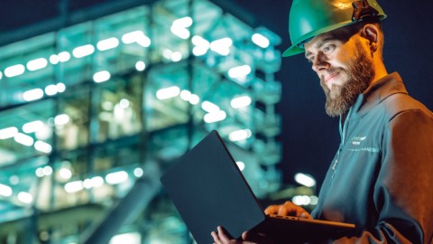 Ein Ingenieur mit grünem Helm arbeitet nachts an einem Laptop vor einem beleuchteten modernen Gebäude.